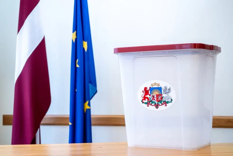 Caurspīdīga vēlēšanu kaste ar sarkanu vāku un Latvijas valsts ģerboni, fonā Latvijas un Eiropas Savienības karogi