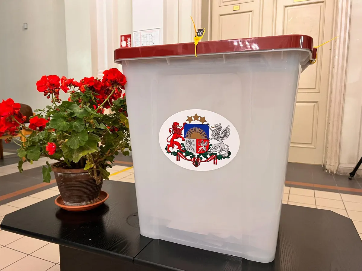 Aizplombēta caurspīdīga vēlēšanu kaste ar Latvijas ģerboni uz galda, blakus puķupods ar sarkanām pelargonijām